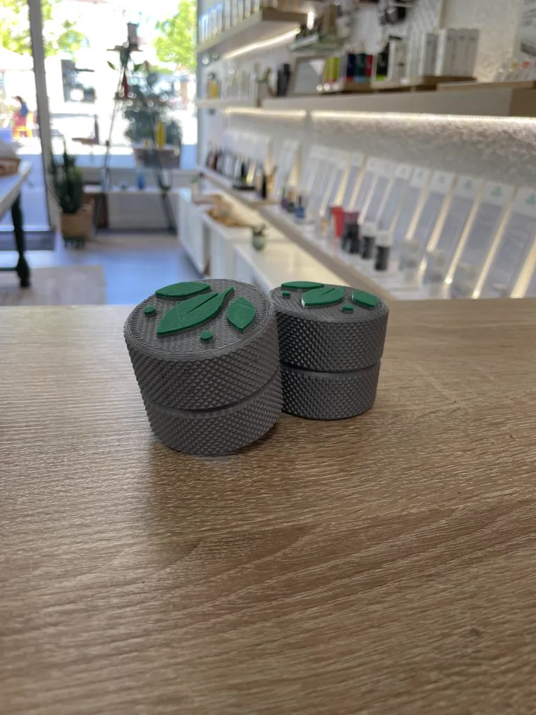 Deux petites boîtes moletées grises au couvercle orné d’un logo vert, posées sur le comptoir en bois d’une boutique