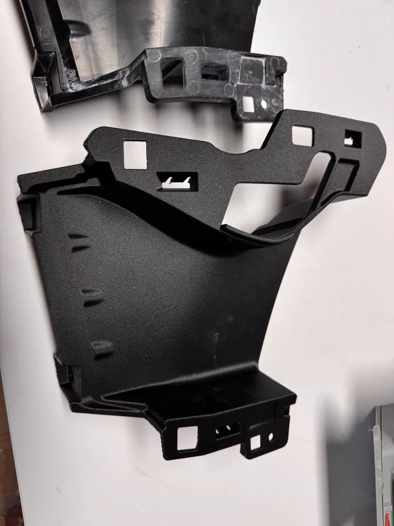Une pièce technique noire de carrosserie automobile, imprimée en 3D, avec ses pattes de fixation, posée sur une table blanche