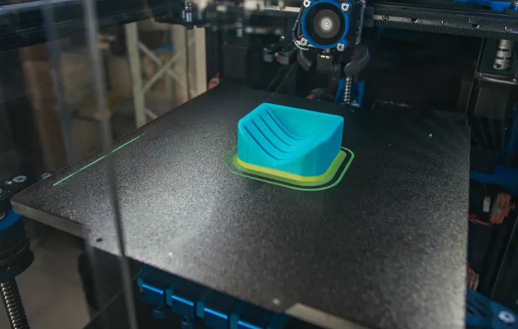 Une pièce turquoise en cours d’impression, posée au milieu d’un plateau noir, sous la tête d’une imprimante 3D